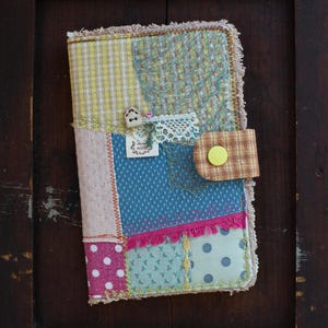 May include: A rectangular fabric journal cover featuring a patchwork design of various patterned textiles in yellow, blue, pink, and green. It includes a brown plaid tab with a yellow snap button, a small wooden house button, a metal key charm, and lace trim.