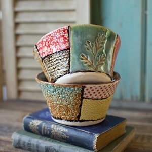 Könnte beinhalten: Zwei Keramikschalen mit Patchwork-Design, übereinander gestapelt. Die Schalen weisen verschiedene Farben und Muster auf, darunter rote Blumen, Grün mit Blatteindrücken und gelbe Punkte. Sie liegen auf antiken Büchern.