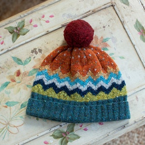 May include: A colorful knitted beanie with a pompom on top. The beanie has a chevron pattern in shades of blue, green, and white, with a solid orange top and a teal brim.