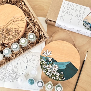 May include: A paint by number kit featuring a round wooden canvas with a mountain and flower design. The kit includes five small bottles of acrylic paint, a paintbrush, and a numbered design sheet.