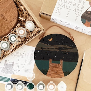 May include: A paint by number kit featuring a round wooden canvas with a nighttime scene of a dock extending into a lake with a crescent moon and stars in the sky. The kit includes six paint pots, a paintbrush, and a wooden hanger.