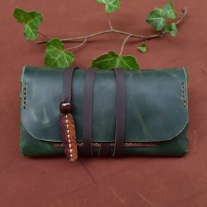 Pode incluir: Uma bolsa de tabaco de couro verde com tiras de couro marrom e um amuleto de pena decorativo.