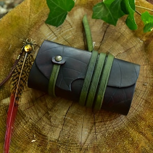 Peut inclure: Un petit carnet en cuir marron foncé avec une sangle en cuir vert et un marque-page en plume. Le carnet est posé sur une surface en bois.
