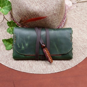 Puede incluir: Una cartera de cuero verde con una correa de cuero marr&oacute;n y un dije de pluma de madera. La cartera est&aacute; encima de un sombrero de paja.