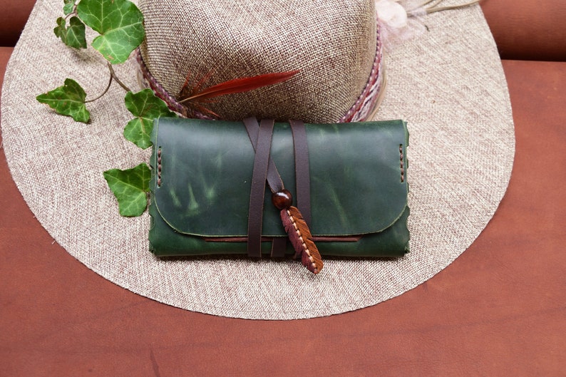 Puede incluir: Una cartera de cuero verde con una correa de cuero marr&oacute;n y un dije de pluma de madera. La cartera est&aacute; encima de un sombrero de paja.