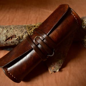 Könnte beinhalten: Eine braune Lederrolle mit einem Riemen und einer Schließe. Die Rolle liegt auf einem Stück Rinde.