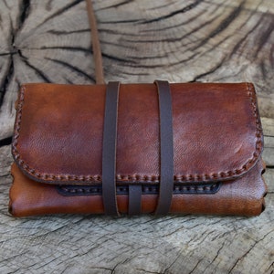 Könnte beinhalten: Eine braune Ledergeldbörse mit zwei Riemen und einem genähten Design. Die Geldbörse ist geschlossen und liegt auf einer Holzoberfläche.