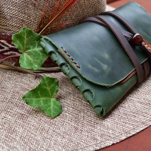 Puede incluir: Una cartera de cuero verde con una correa de cuero marr&oacute;n y cierre de hebilla. La cartera est&aacute; sobre un sombrero de paja tejido.