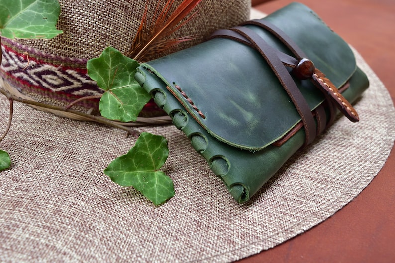 Puede incluir: Una cartera de cuero verde con una correa de cuero marr&oacute;n y cierre de hebilla. La cartera est&aacute; sobre un sombrero de paja tejido.