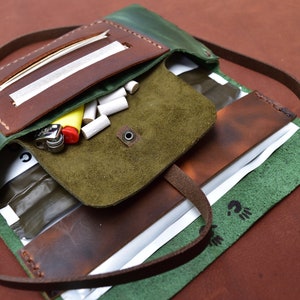 Puede incluir: Una bolsa enrollable de cuero verde y marr&oacute;n con una correa. La bolsa tiene varios compartimentos y est&aacute; abierta para revelar un encendedor y puntas de filtro.