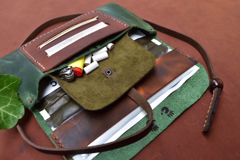Puede incluir: Una bolsa enrollable de cuero verde y marr&oacute;n con una correa. La bolsa tiene varios compartimentos y est&aacute; abierta para revelar un encendedor y puntas de filtro.