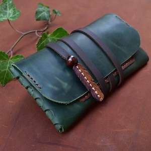 Puede incluir: Una cartera de cuero verde con una correa de cuero marr&oacute;n y un adorno de pluma decorativo.