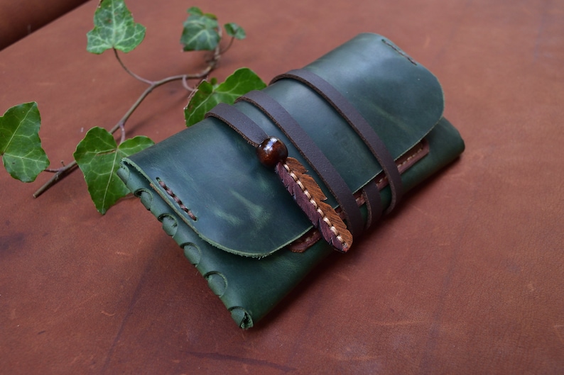 Puede incluir: Una cartera de cuero verde con una correa de cuero marr&oacute;n y un adorno de pluma decorativo.