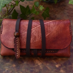 Peut inclure: Un portefeuille en cuir marron avec un motif de feuille et une sangle en cuir marron. Le portefeuille a un pompon en cuir marron avec une perle en bois.