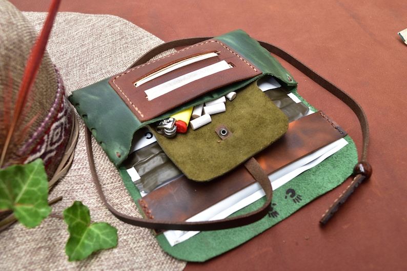 Puede incluir: Una bolsa de tabaco de liar de cuero verde con una solapa de cuero marr&oacute;n y una correa. La bolsa est&aacute; abierta y contiene papeles de liar, filtros y un encendedor.