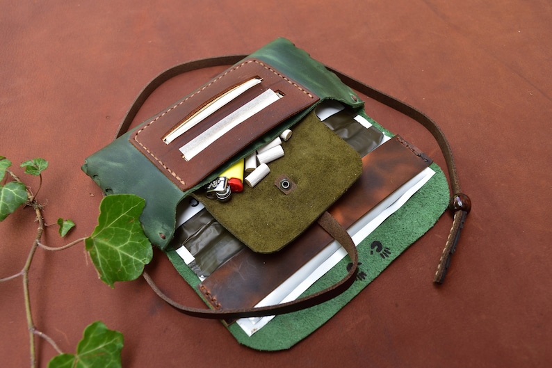 Puede incluir: Un estuche enrollable de cuero verde con una solapa de cuero marr&oacute;n y una correa. El estuche est&aacute; abierto y contiene un encendedor, filtros y papeles de liar.
