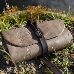May include: A brown leather roll-up pouch with a strap and buckle closure. The pouch is lying on a bed of green grass and brown soil.