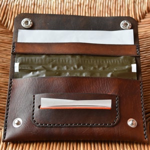 Op de afbeelding: Een bruine leren tabakspouch met een klep en twee drukknopen. De pouch is gevoerd met wit papier en heeft een groene voering aan de binnenkant. Er is een klein vakje aan de voorkant van de pouch met een witte papieren voering en een oranje rand.