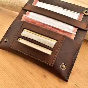 Könnte beinhalten: Ein braunes Leder-Rolltablett mit zwei Schlitzen für Rollpapiere. Das Tablett hat Messingakzente und Nähte.