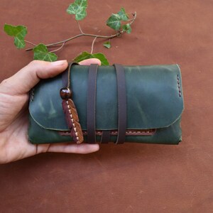 Puede incluir: Una cartera de cuero verde con correas marrones y un borla de cuero marr&oacute;n. La cartera se sostiene en una mano.