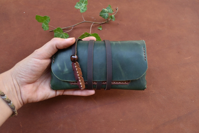 Puede incluir: Una cartera de cuero verde con correas marrones y un borla de cuero marr&oacute;n. La cartera se sostiene en una mano.