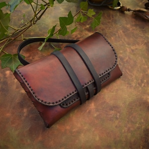 Peut inclure: Une pochette en cuir marron avec des lanières et des coutures en cuir noir. La pochette est fermée et posée sur une surface marron.