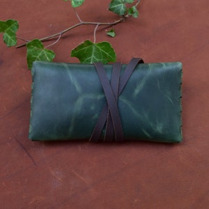 Puede incluir: Una bolsa de cuero verde con correas de cuero marr&oacute;n atadas alrededor. La bolsa est&aacute; sobre una superficie de cuero marr&oacute;n.