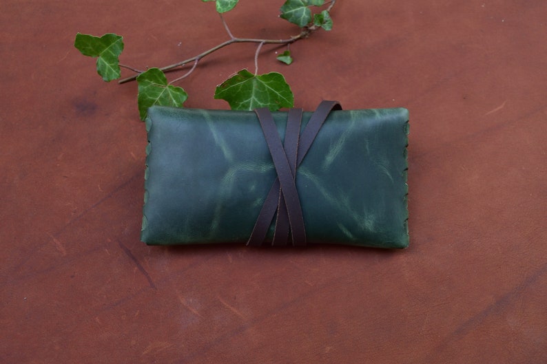 Puede incluir: Una bolsa de cuero verde con correas de cuero marr&oacute;n atadas alrededor. La bolsa est&aacute; sobre una superficie de cuero marr&oacute;n.