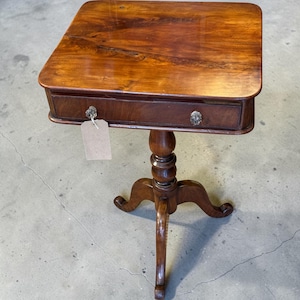 May include: A square wooden side table with a polished brown finish, featuring a single drawer with two clear glass knobs. The table is supported by a central pedestal base that branches into three curved legs. A blank beige tag hangs from one knob.