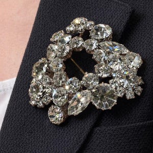 May include: A dazzling brooch featuring numerous clear crystals set in a silver-toned metal. The brooch is affixed to a dark navy blazer, providing a striking contrast. The crystals are arranged in a clustered, floral-like pattern.