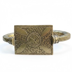 May include: A brass-toned metal cuff bracelet featuring a rectangular centrepiece with an etched geometric pattern of circles and lines. The band has a ribbed texture and connects to the central plate with small, decorative circular accents.