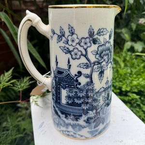 May include: Cream-coloured pitcher with a blue floral and pagoda design. The pitcher has a gold rim and handle. The handle is curved and has a decorative element at the top. The pitcher is sitting on a white surface.