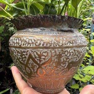 May include: A round, metallic planter with a scalloped rim and a tapered base. The exterior features intricate, etched floral and animal motifs in a dark, weathered bronze finish with contrasting light-coloured recessed details throughout the surface.
