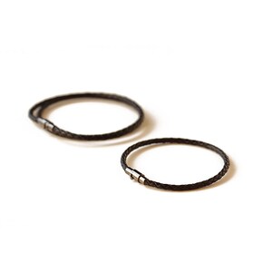 May include: Two black braided leather bracelets with silver-colored clasps are displayed against a white background. The bracelets are circular and appear to be for the wrist.