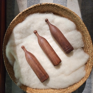 May include: Three smooth, brown wooden darning eggs with elongated handles, arranged diagonally on a bed of soft, off-white wool fiber inside a round, woven wicker basket. Each wooden tool features a rounded top and a base with etched lines.