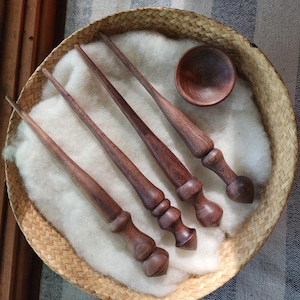 May include: Four dark brown wooden drop spindles with tapered shafts and decorative turned handles, arranged diagonally alongside a small matching wooden bowl, all resting on a bed of white wool inside a round woven basket.