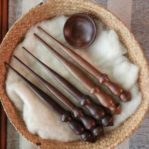 May include: A set of four dark brown wooden drop spindles with tapered shafts and rounded handles, accompanied by a small matching wooden bowl, all arranged on a bed of white wool inside a round woven basket with a textured rim.