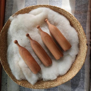 May include: Four light brown wooden darning eggs with elongated, bottle-like shapes and smooth, rounded handles. These wooden tools are arranged in a row, resting on a soft, white, fluffy layer of raw wool inside a round, woven straw basket.