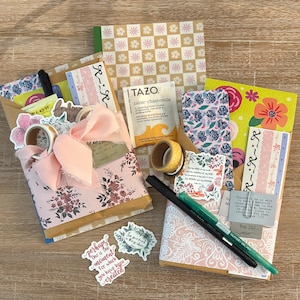 K&ouml;nnte beinhalten: Eine Sammlung von Schreibwaren, darunter dekoratives Papier, Aufkleber, Washi-Tape, Stifte und ein Teebeutel. Die Artikel sind auf einer Holzoberfl&auml;che angeordnet, mit floralen und gemusterten Designs in Rosa, Gelb und Blau.