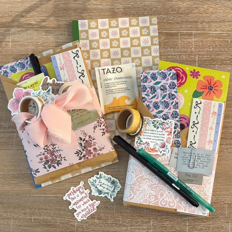 K&ouml;nnte beinhalten: Eine Sammlung von Schreibwaren, darunter dekoratives Papier, Aufkleber, Washi-Tape, Stifte und ein Teebeutel. Die Artikel sind auf einer Holzoberfl&auml;che angeordnet, mit floralen und gemusterten Designs in Rosa, Gelb und Blau.