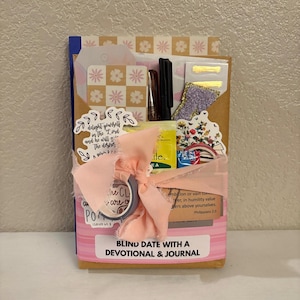 K&ouml;nnte beinhalten: Ein Geschenkset mit der Aufschrift "BLIND DATE WITH A DEVOTIONAL & JOURNAL". Das Set enth&auml;lt ein Notizbuch, Aufkleber, Stifte und andere Schreibwaren. Es ist mit einem rosa Band und dekorativem Klebeband umwickelt.