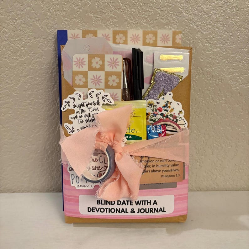 K&ouml;nnte beinhalten: Ein Geschenkset mit der Aufschrift "BLIND DATE WITH A DEVOTIONAL & JOURNAL". Das Set enth&auml;lt ein Notizbuch, Aufkleber, Stifte und andere Schreibwaren. Es ist mit einem rosa Band und dekorativem Klebeband umwickelt.