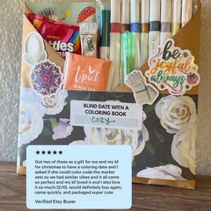 May include: A gift box with a floral design, containing markers, candy, tea, and stickers. The box includes the text "Blind Date With A Coloring Book Cozy" and the words "be joyful always".