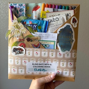 May include: A brown paper package labeled "Blind Date with a Coloring Book - Floral" filled with various items. Includes markers, tea, candy, stickers, and a cassette tape. Decorated with floral and mushroom-themed stickers and patterns.