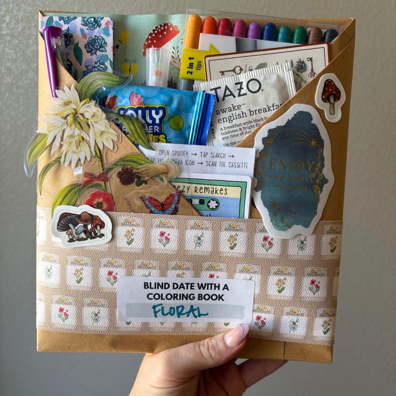 May include: A brown paper package labeled "Blind Date with a Coloring Book - Floral" filled with various items. Includes markers, tea, candy, stickers, and a cassette tape. Decorated with floral and mushroom-themed stickers and patterns.
