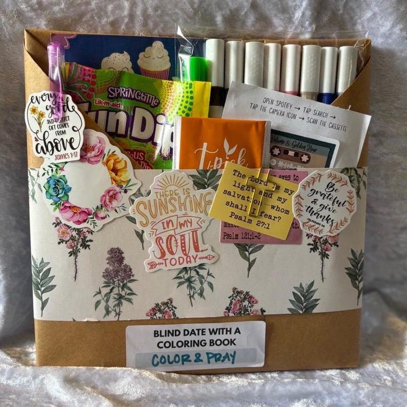 K&ouml;nnte beinhalten: Eine Geschenkbox mit floral gemusterter Verpackung, die Snacks, Marker und Aufkleber enth&auml;lt. Die Box enth&auml;lt den Text "Blind Date with a Coloring Book" und "Color & Pray". Dekorative Aufkleber mit inspirierenden Zitaten sind ebenfalls sichtbar.
