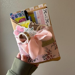 May include: A decorative gift box wrapped in floral and patterned paper, tied with a pink ribbon. The box contains stickers, pens, and washi tape. The box has the text "In Christ Alone my hope" and "For when I'm afraid I will trust in you".