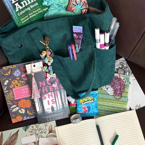 May include: A green corduroy tote bag filled with books, pens, and other items. The bag contains a coloring book, a novel titled "Nowhere She Is", and a Sudoku puzzle book. Other items include pens, a tube of lotion, and a bag of candy.