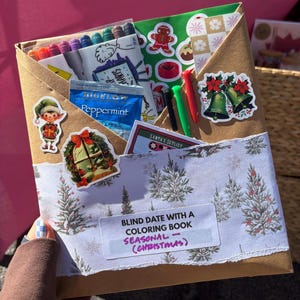 May include: A brown paper envelope with a Christmas theme, filled with coloring supplies and stickers. The envelope contains colored pencils, stickers of bells, a gingerbread man, and a tea bag. Text on the envelope reads "Blind Date with a Coloring Book - Seasonal (Christmas)".