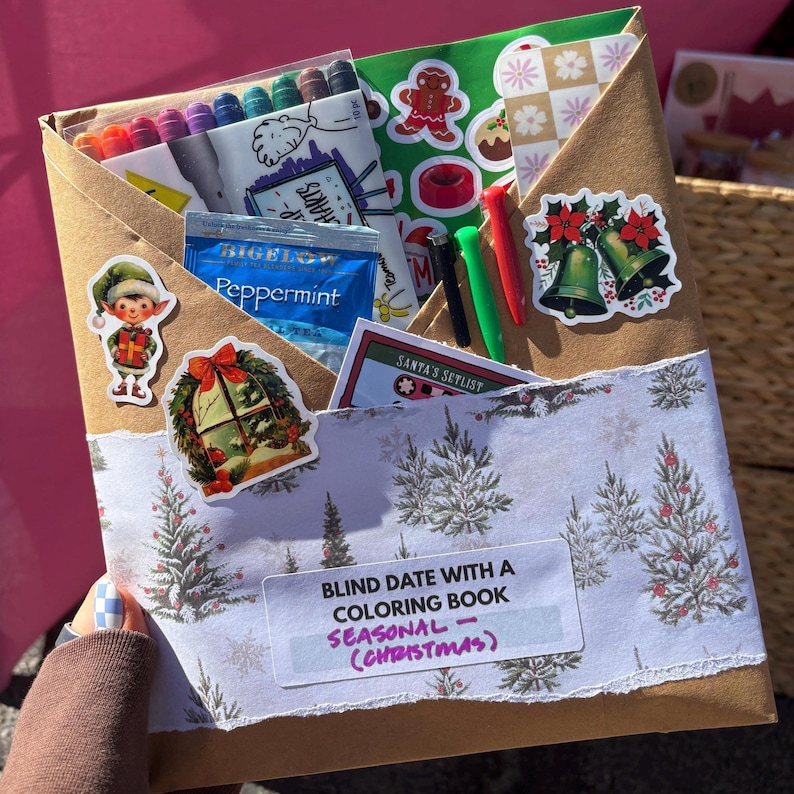 May include: A brown paper envelope with a Christmas theme, filled with coloring supplies and stickers. The envelope contains colored pencils, stickers of bells, a gingerbread man, and a tea bag. Text on the envelope reads "Blind Date with a Coloring Book - Seasonal (Christmas)".